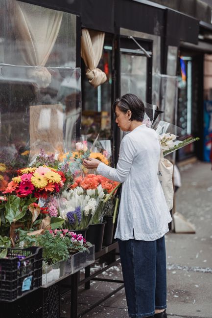 A woman with short black hair, dressed in a white long-sleeved top and dark blue jeans, stands outdoors in front of a florist shop window. She is carefully selecting and arranging vibrant fresh flower