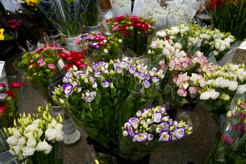 A vibrant display of fresh flowers arranged in bouquets on a floral shop counter, featuring purple and white lisianthus, white roses, pink ranunculus, and red and pink daisies. The flowers are wrapped