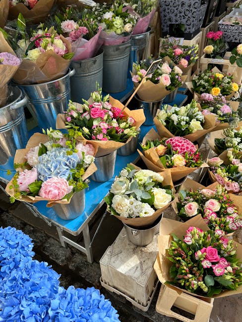 A display of fresh floral bouquets arranged in small metal buckets and wrapped in brown craft paper, featuring a variety of flowers including pink roses, white lisianthus, blue hydrangeas, and light g