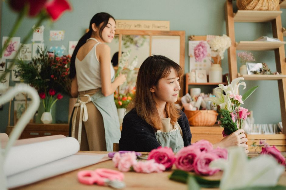 A young woman with light brown hair and a focused expression is seated at a wooden table inside a flower shop, holding a small floral arrangement featuring white lilies and pink roses with green folia