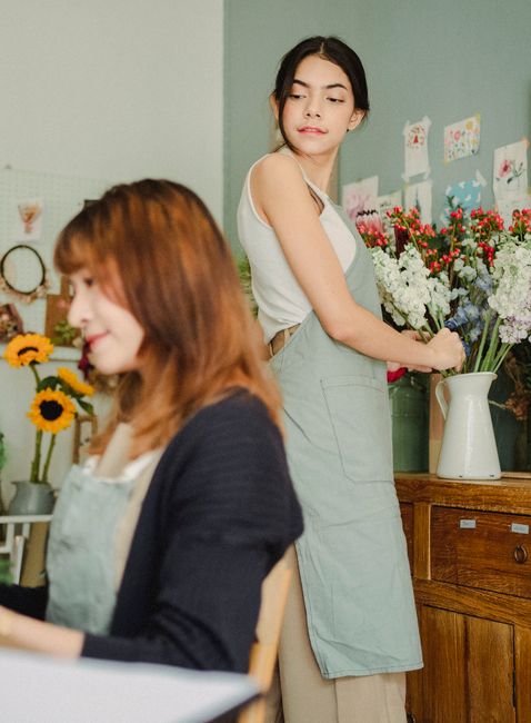 A woman arranging a vibrant bouquet of fresh flowers on a wooden counter inside a florist shop. The bouquet features an assortment of blooms in red, white, purple, and pink hues, with green foliage pr