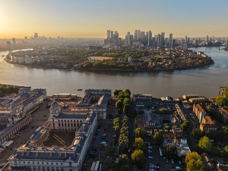 Aerial view of the London skyline at sunset, featuring the River Thames meandering through the city. In the foreground, there are historic white and grey neoclassical buildings with domed rooftops, su
