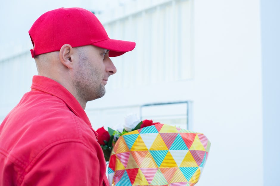 A delivery person wearing a red cap and matching red jacket holds a large, colorful, geometric-patterned gift box wrapped with yellow, red, blue, and green triangles. The person's profile shows a focu