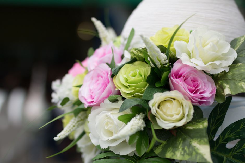 A floral arrangement featuring soft pastel-colored roses and peonies in shades of white, cream, pink, and light green, set against a blurred indoor background. The bouquet is composed of fresh, velvet